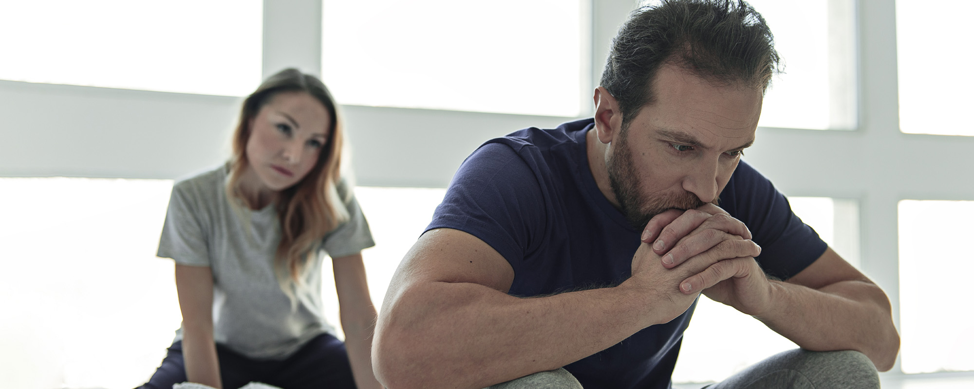 frustrated husband on bed with anxious looking wife behind him